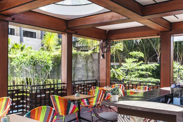 A covered open-air dining area with colourful woven chairs, wooden beams, a ceiling fan and a bar counter, looking out onto a garden with palm and frangipani trees.