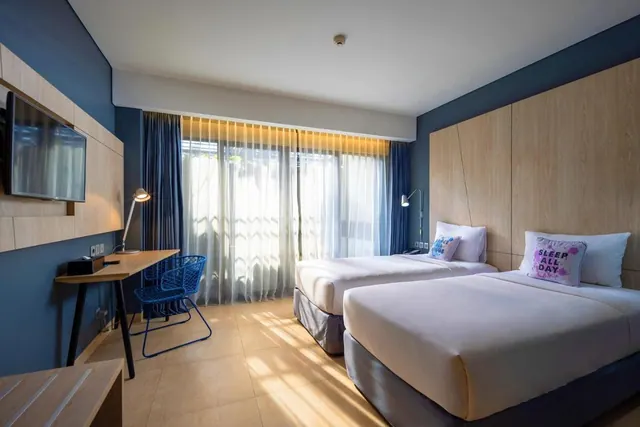 A bedroom with two single beds, a wall-mounted television, a wooden desk with a blue chair, and sheer curtains over a large window.