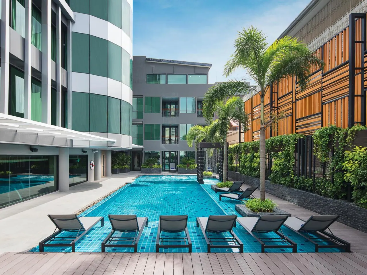An outdoor lap pool with blue tiles and grey sun loungers along its edge, with palm trees and a green wall beside it.