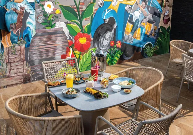 An outdoor dining table with drinks and dishes sits in front of a colourful painted mural of a tuk-tuk and plants, with rattan chairs around it.