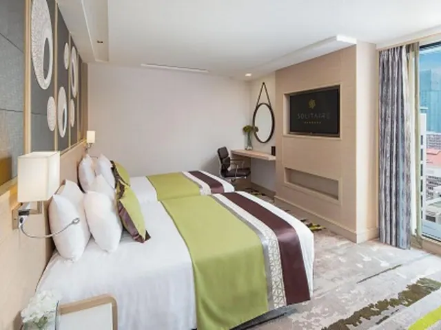 A hotel bedroom with two beds, a wall-mounted television, a desk with a chair, and a window showing city buildings outside.