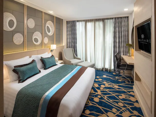 A hotel bedroom with a king bed, an armchair with ottoman, a desk with chair, and a wall-mounted television beside tall curtained windows.