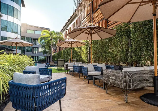 An outdoor terrace with blue and grey wicker seating under beige parasols, backed by a tall green hedge and a wooden-slatted building.