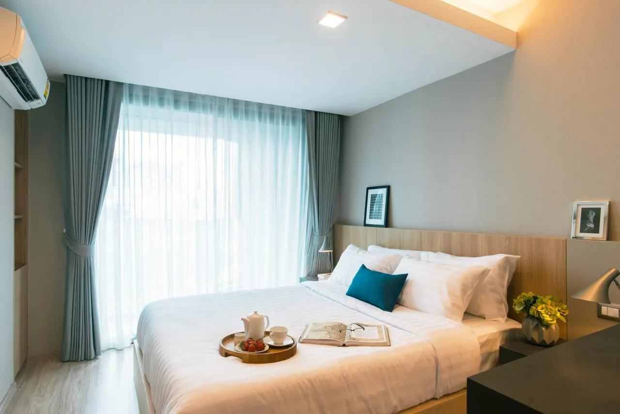A made double bed with white linen and a teal cushion, with a tray of teapot, cups and fruit and an open book, beside a large window with sheer curtains.