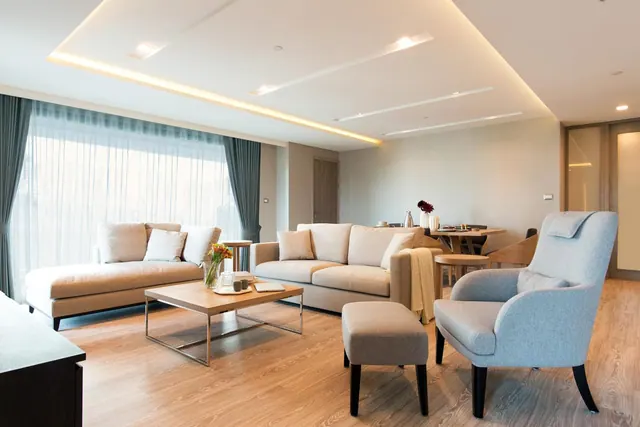 A staged living room with beige sofas, a wooden coffee table, a grey armchair and a dining table in the background, beside a large window covered with sheer curtains.