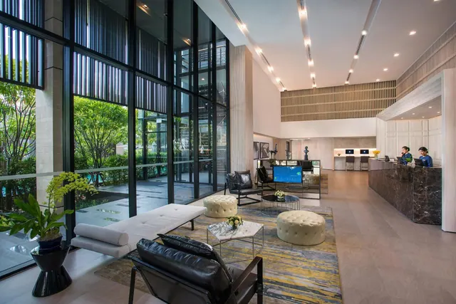 The lobby has a lounge seating area with leather armchairs and ottomans, floor-to-ceiling windows overlooking greenery, and a reception desk where two staff are working.