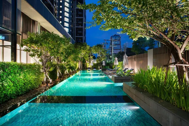 A long illuminated lap pool with a turquoise tiled floor runs between tall trees, sun loungers and closed umbrellas beside a residential tower at dusk.