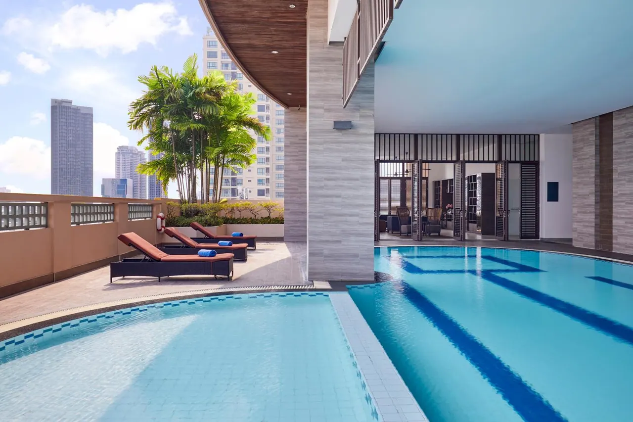 An outdoor lap pool with blue lane lines sits beside a terrace of sun loungers, with city towers visible beyond the railing.