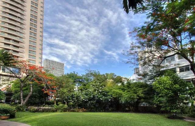 A trimmed lawn with flowering trees and shrubs, with tall residential towers visible behind under a partly cloudy sky.