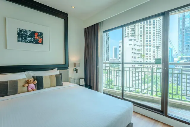 A bedroom with a made king bed and a teddy bear on the pillows, with floor-to-ceiling windows opening onto a balcony overlooking high-rise buildings.