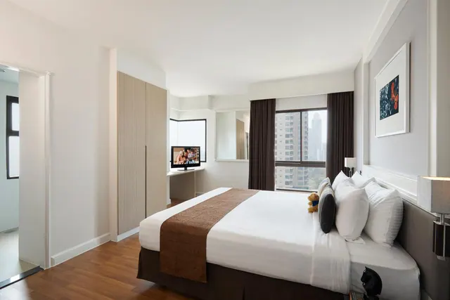 A king bed with white linens and a brown runner sits in a bedroom beside a wardrobe, a wall-mounted television and a window overlooking high-rise buildings.