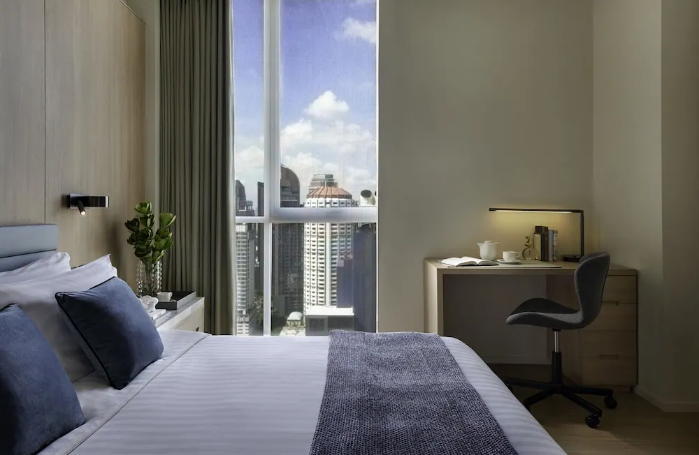 A bedroom with a made king bed facing a tall window that looks out over high-rise buildings under a blue sky, with a desk and chair to the right.