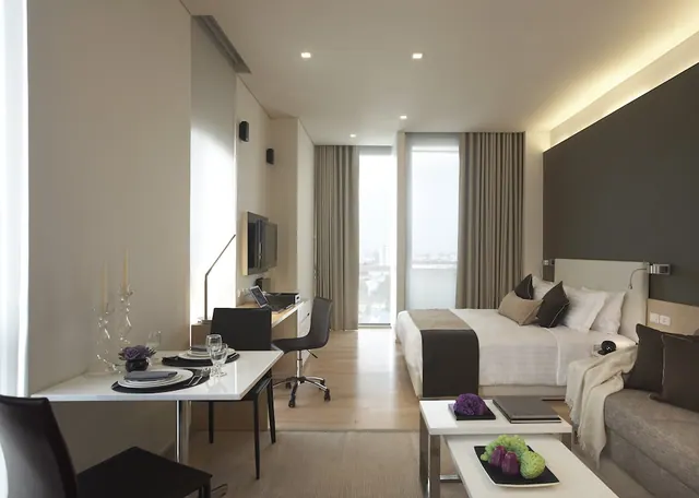 A studio room with a double bed, sofa, dining table and work desk, with floor-to-ceiling windows looking out onto city buildings.