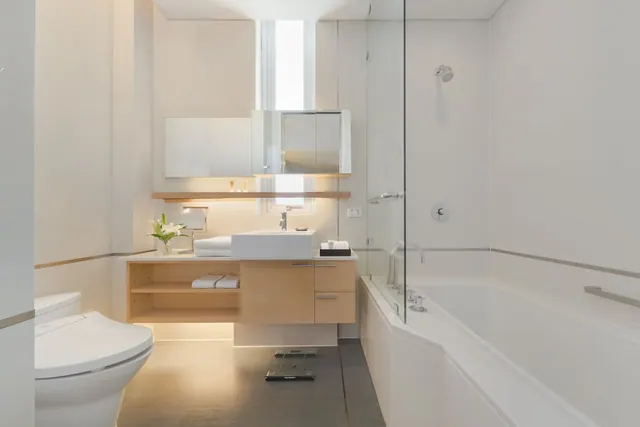 A bathroom with a wall-mounted wooden vanity and single basin, a toilet, a bathtub beside a glass shower screen with an overhead shower head, and folded towels on a shelf.