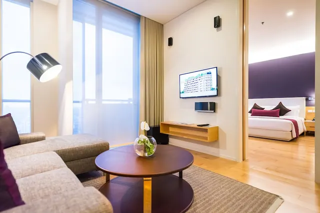 A living area with a grey sofa, a round wooden coffee table and a wall-mounted television, with a bedroom with a purple accent wall visible through the doorway.