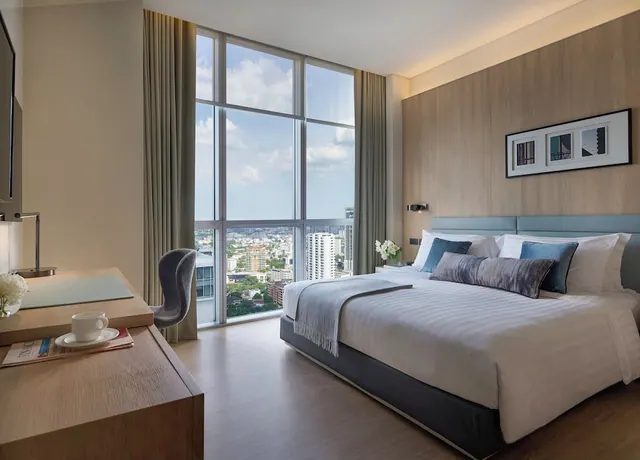 A bedroom with a king bed, a wooden desk with a cup and saucer, and floor-to-ceiling windows looking out over city buildings.