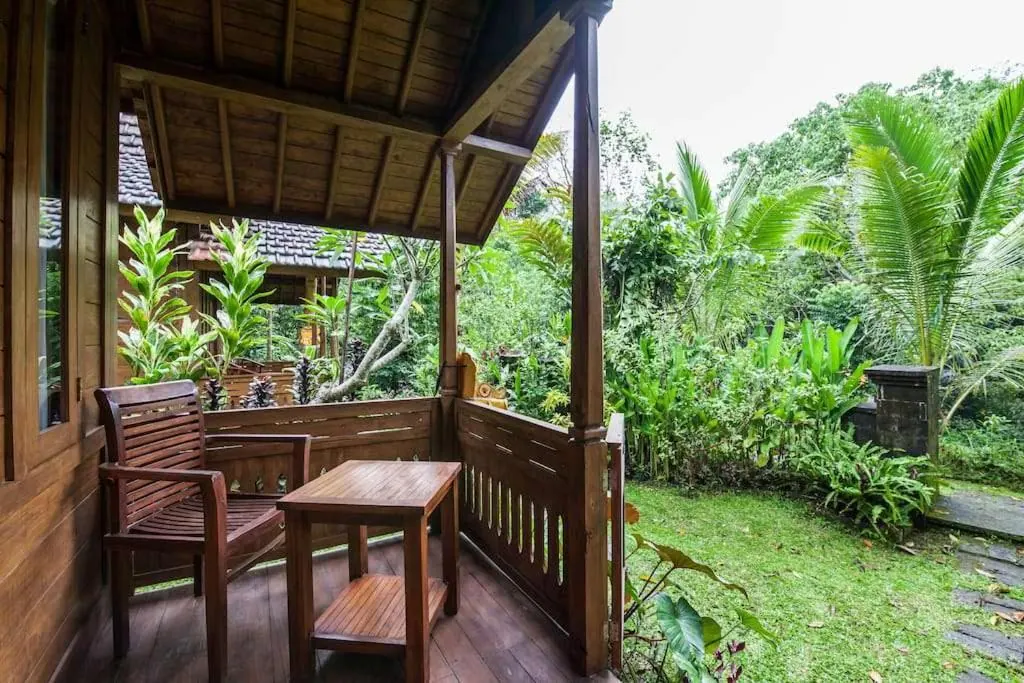 A wooden veranda with a wooden chair and side table overlooks a lawn and lush tropical garden with palm trees.