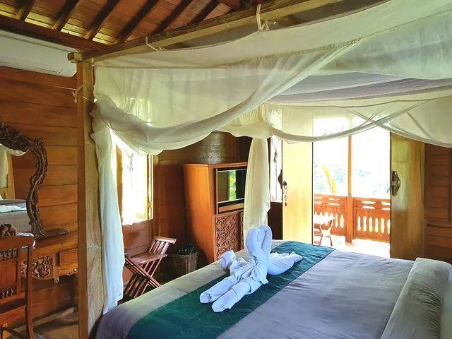 A bedroom with a canopy mosquito net over a bed with a folded towel animal, wooden walls, and an open doorway to a balcony with greenery beyond.