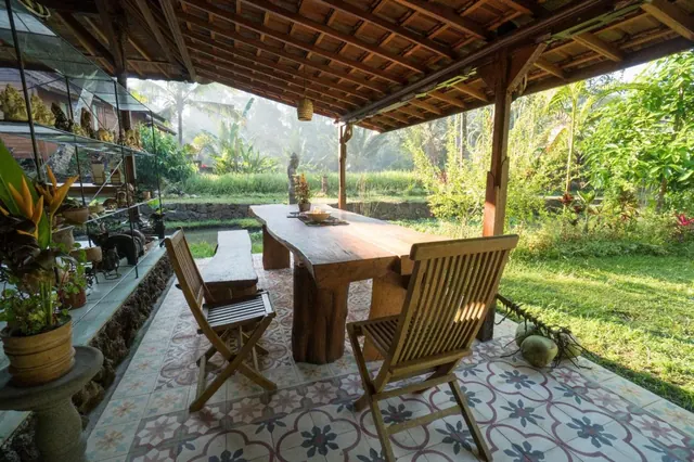 A covered open-air terrace with a wooden dining table, folding teak chairs and patterned floor tiles, overlooking a green garden and rice field.