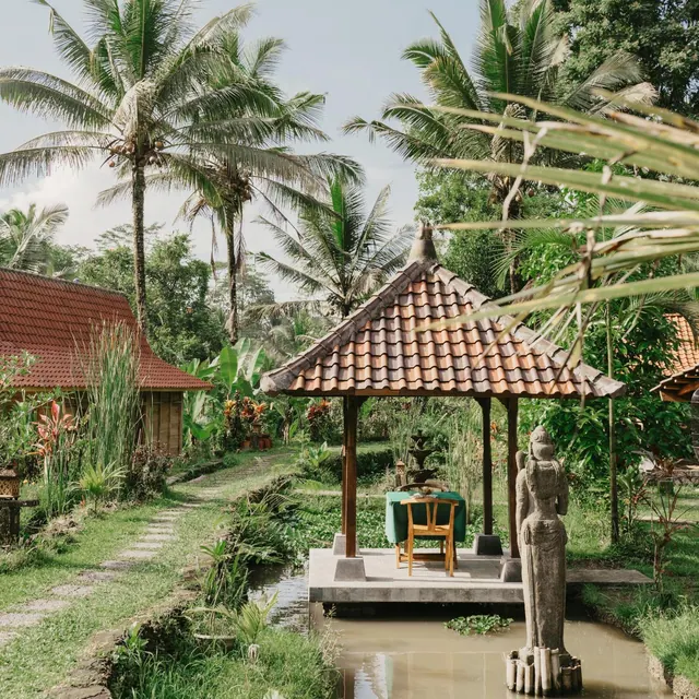 A garden pavilion with a table and chair stands over a water channel, beside a stone statue and palm trees.