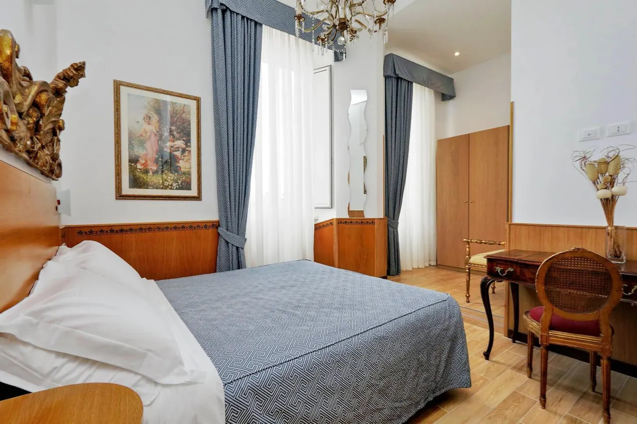 A bedroom with a double bed under a blue patterned cover, a wooden wardrobe, a writing desk with a cane chair, and a framed painting on the wall.