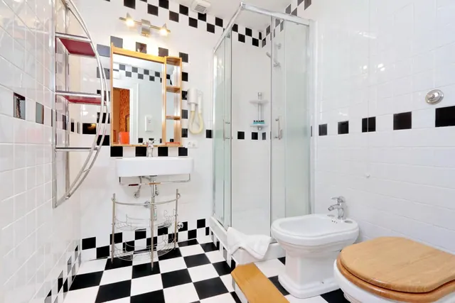 A bathroom with black and white checkered floor and wall tiles, a glass-enclosed shower, a pedestal sink, a bidet and a toilet with a wooden seat.