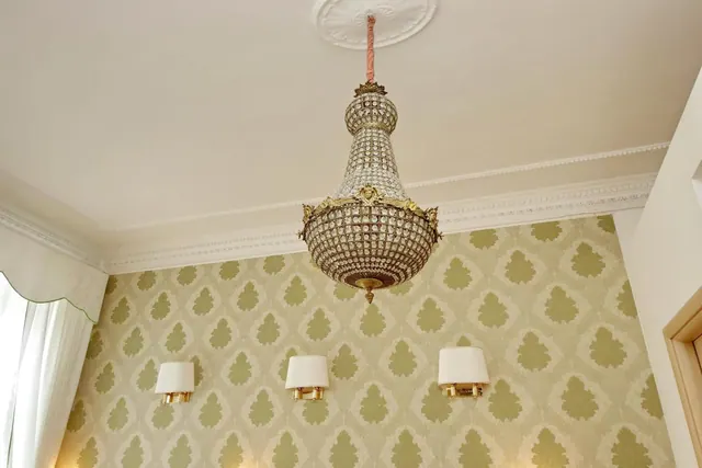 A crystal chandelier hangs from a white ceiling with decorative moulding above green patterned wallpaper, with three wall sconces and a curtain at the left.