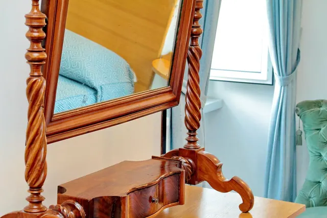 A wooden swivel dressing table with a barley-twist frame and a small serpentine drawer stands beside a window with blue curtains and a green tufted armchair.