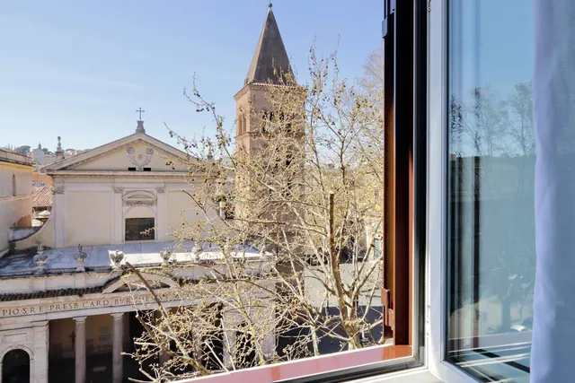 An open window frames a bare tree in front of a church with a tall brick bell tower and a columned building with a pediment under a clear blue sky.