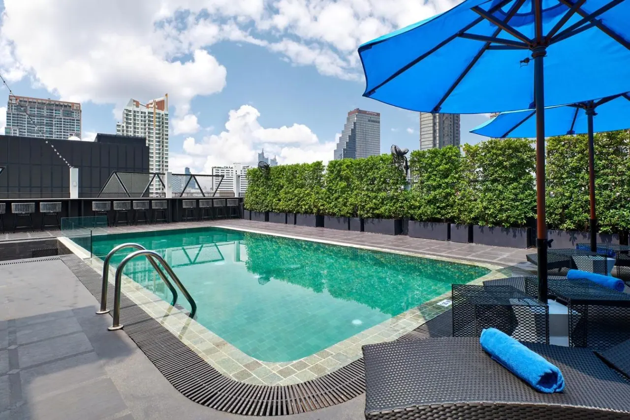 A rooftop swimming pool with blue umbrellas and sun loungers, with city high-rises visible beyond the hedges.