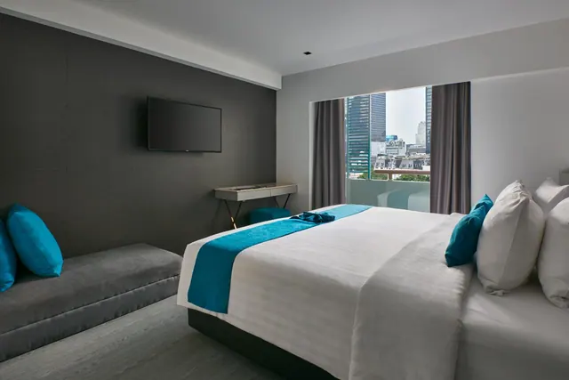 A bedroom with a large bed dressed in white and teal linen, a grey bench with teal cushions, a wall-mounted television, and a window overlooking city skyscrapers.