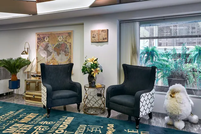 A lobby lounge with two dark wingback armchairs, a framed world map on the wall, a flower arrangement on a side table, and a window looking out onto plants and buildings.