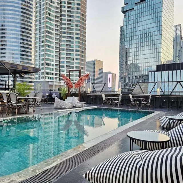 A rooftop swimming pool with striped loungers and tables, with high-rise towers rising behind it.