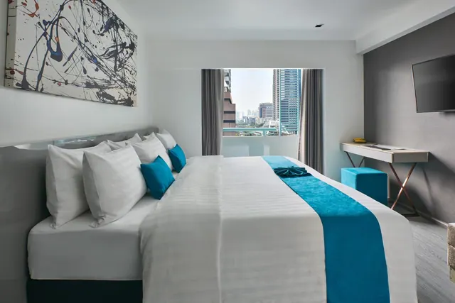 A bedroom with a made king bed with teal accents, a wall-mounted television and a desk, with a window showing city high-rise buildings.