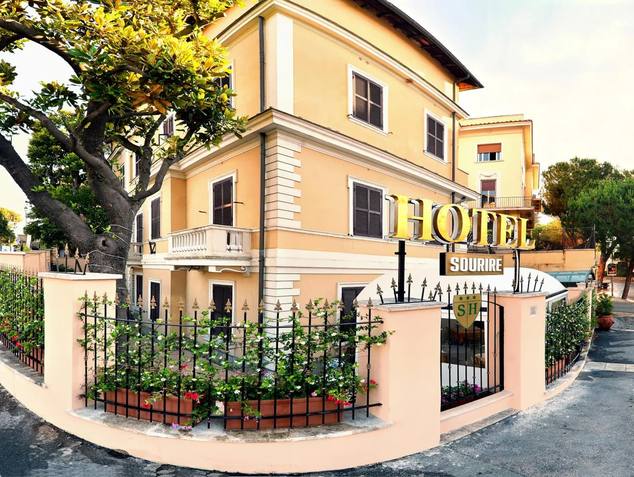 Front exterior of Hotel Sourire, a yellow building behind a black iron fence with flowering plants and a gold hotel sign.