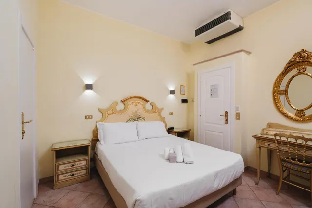 A double bed with a painted wooden headboard, two bedside cabinets, a gilt mirror and a small desk with a chair in a bedroom with a wall-mounted air conditioner.