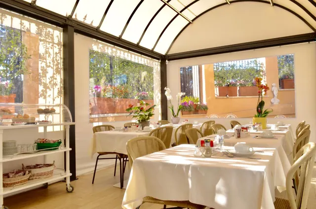 A glass-roofed breakfast room with white-clothed tables and wicker chairs, with windows onto a plant-filled courtyard and a breakfast cart at left.
