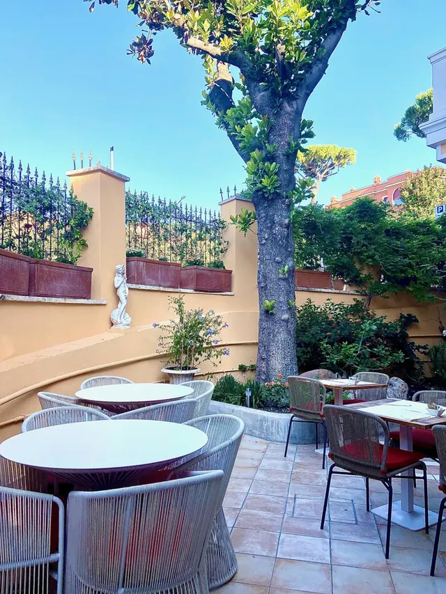 An empty outdoor terrace with round tables and grey woven chairs under a large tree, with a white statue and planters against ochre walls.