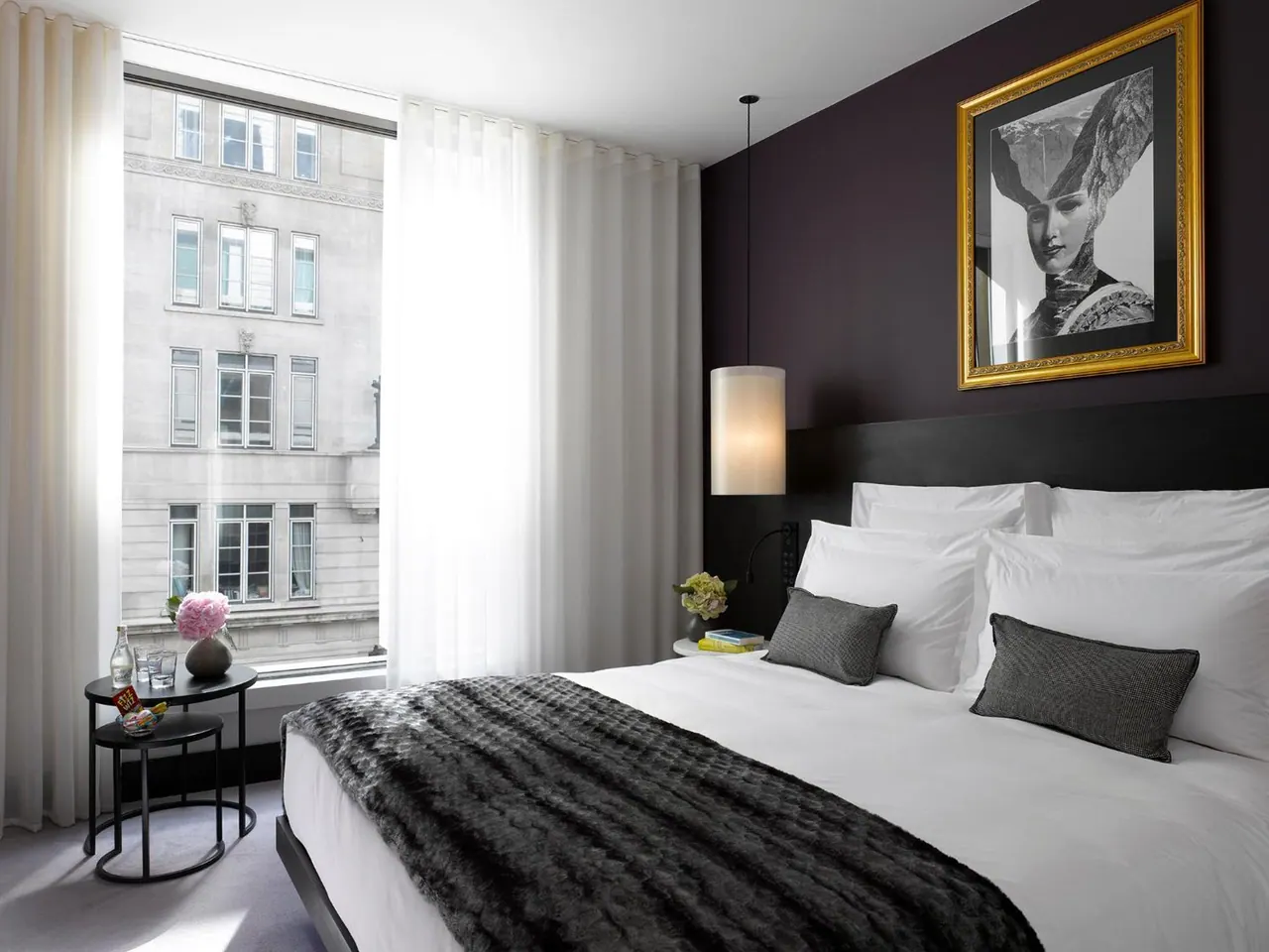 A made king bed with white linen and grey cushions sits beside a tall window with sheer curtains that looks onto a stone building, with a framed black-and-white portrait on the plum-coloured wall above the bed.