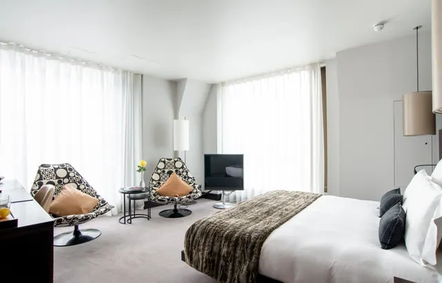 A bedroom with a large bed, two patterned armchairs, a wall-mounted television and tall windows with sheer curtains.