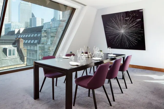 A set dining table with purple upholstered chairs stands in a room with a large angled window overlooking city towers and rooftops.