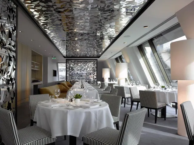 A dining room with white-clothed tables set with glassware, striped upholstered chairs, a mirrored patterned ceiling and large angled windows.