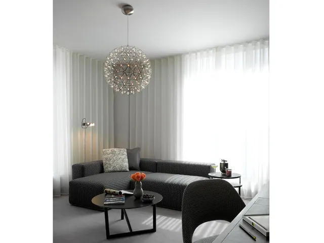 A living room with a spherical pendant light, a grey sofa, a round black coffee table with a vase of orange roses, and sheer curtains over a large window.