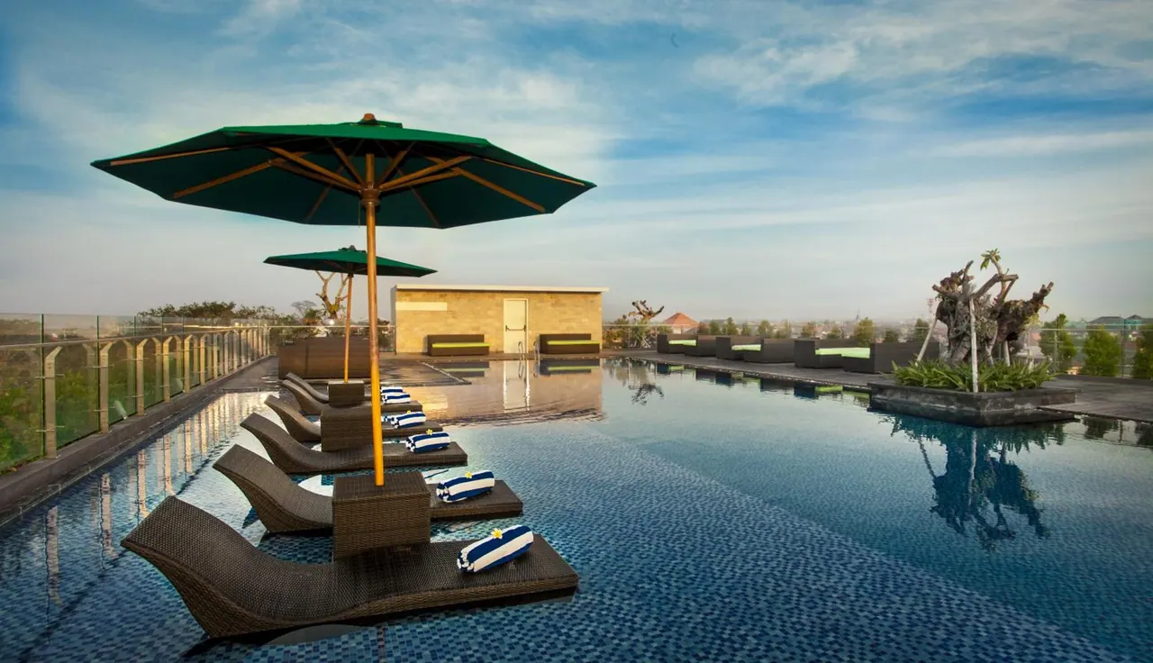 A rooftop swimming pool with sun loungers, green umbrellas and striped towels, with trees and open sky beyond.