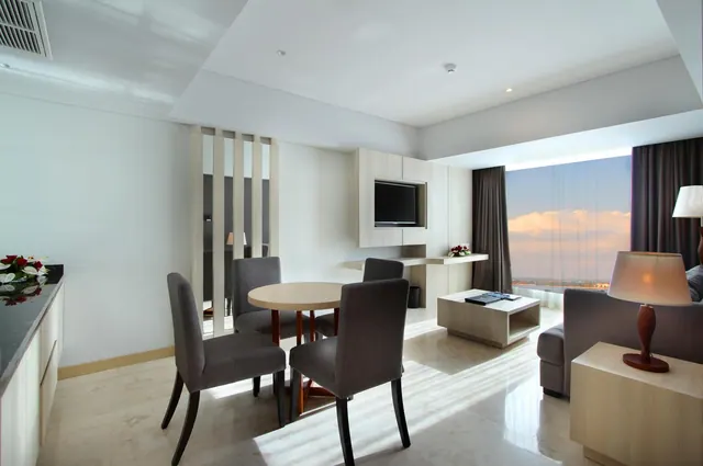 A staged living and dining area in a hotel suite with a round dining table, grey chairs, a wall-mounted television, and large windows showing a sunset sky over distant land.