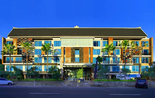 The front facade of a multi-storey hotel with a dark pitched roof, palm trees, and a street with parked cars in the foreground.