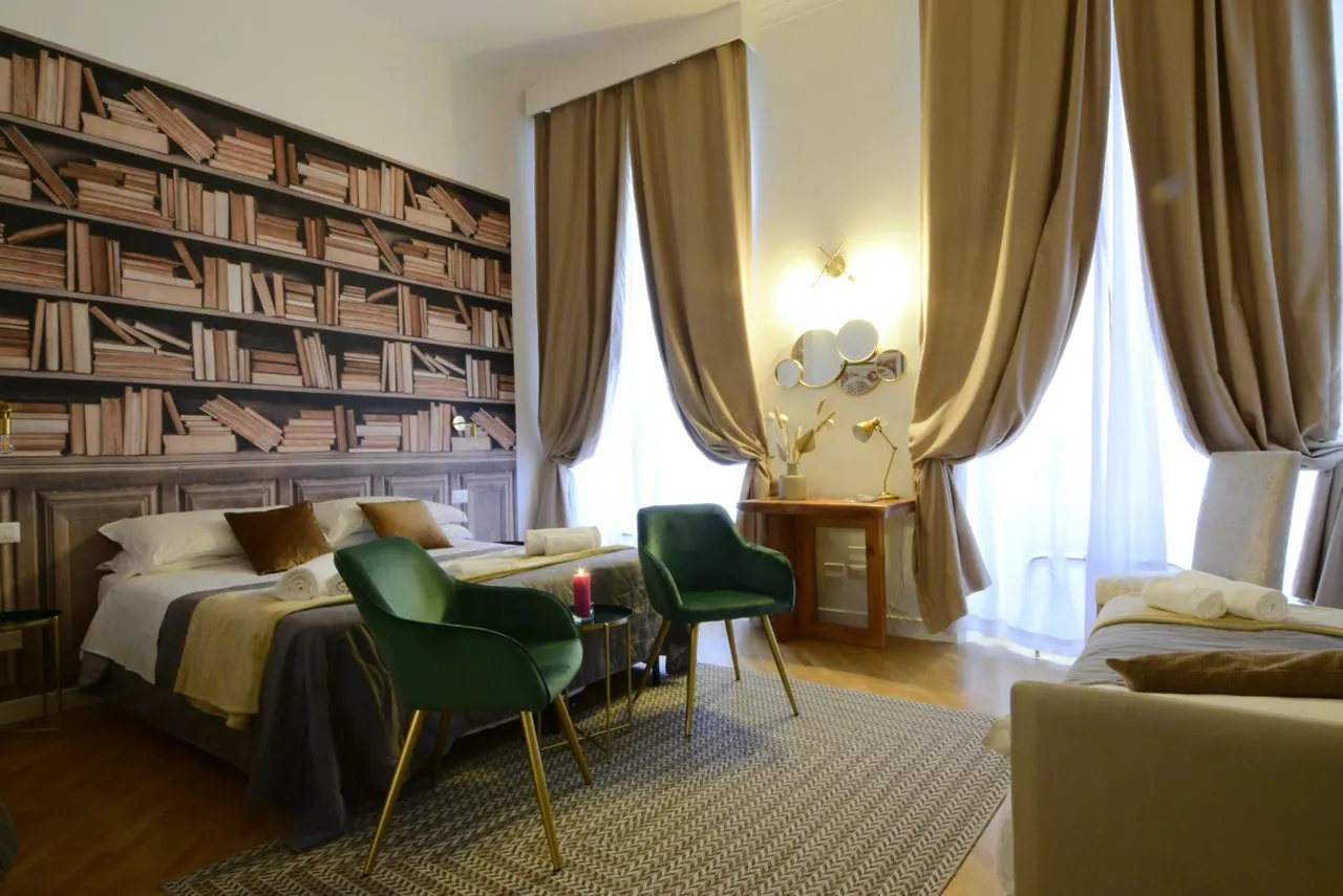 A bedroom with a double bed in front of a bookshelf-patterned wall, two green velvet chairs, a second bed at the right edge, and tall curtained windows.
