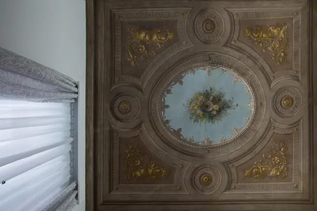 A coffered ceiling with gold ornaments and a painted floral medallion, beside a window with a patterned roman blind.