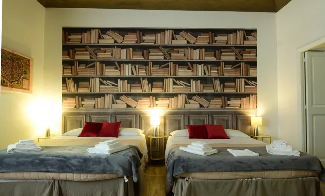 Two double beds with grey throws and folded white towels sit in front of a bookshelf-patterned wall with red cushions and bedside lamps.