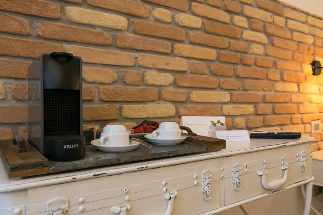 A black Krups coffee machine and two white cups on saucers sit on a tray atop a white distressed chest beside a brick wall.